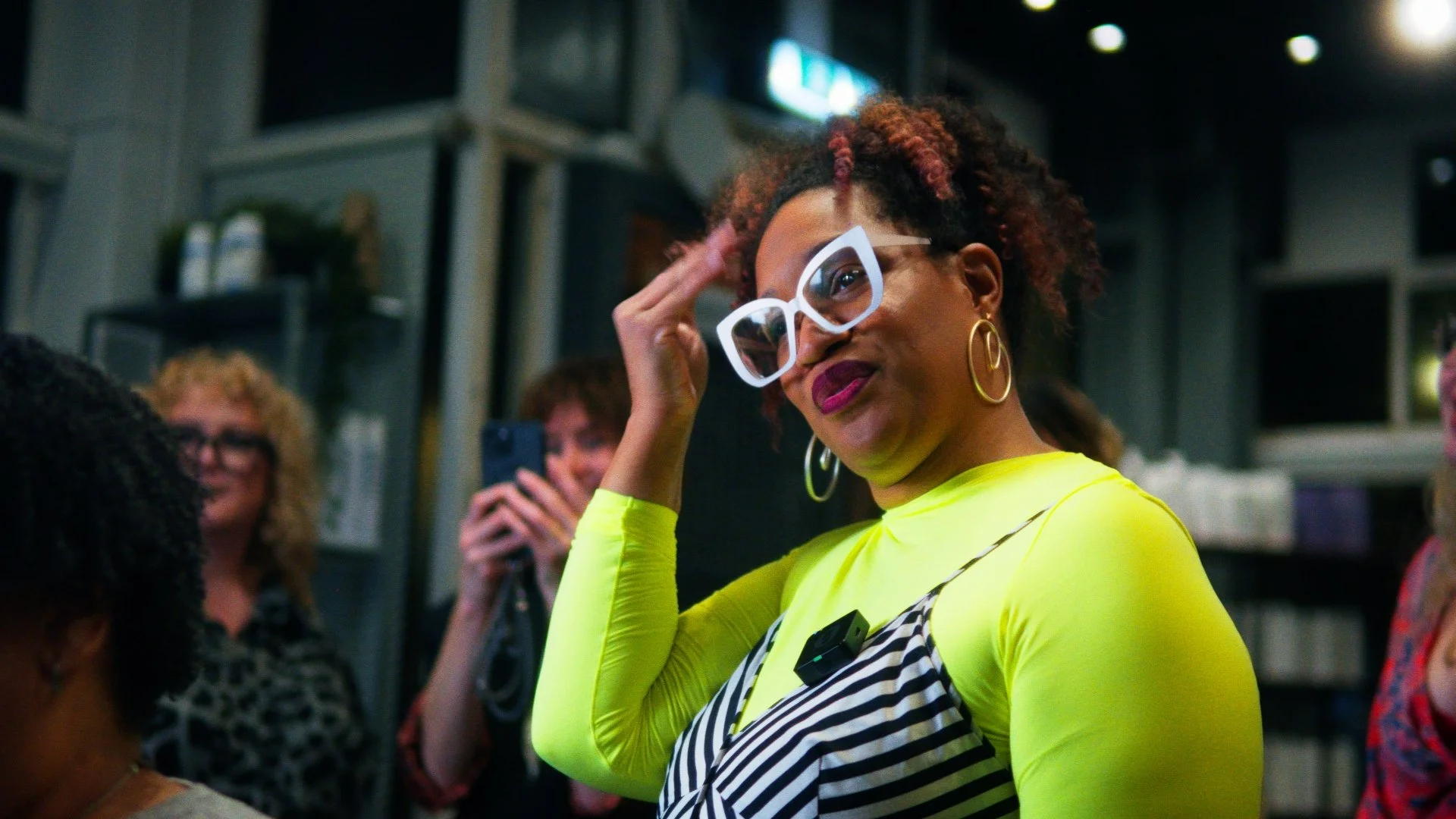Atlanta tight artist Aeleise Jana  wearing large white glasses, bright yellow top, and a striped apron, stands in a room with shelves, talking and touching her head. Others are in the background, one person taking a photo and another smiling.