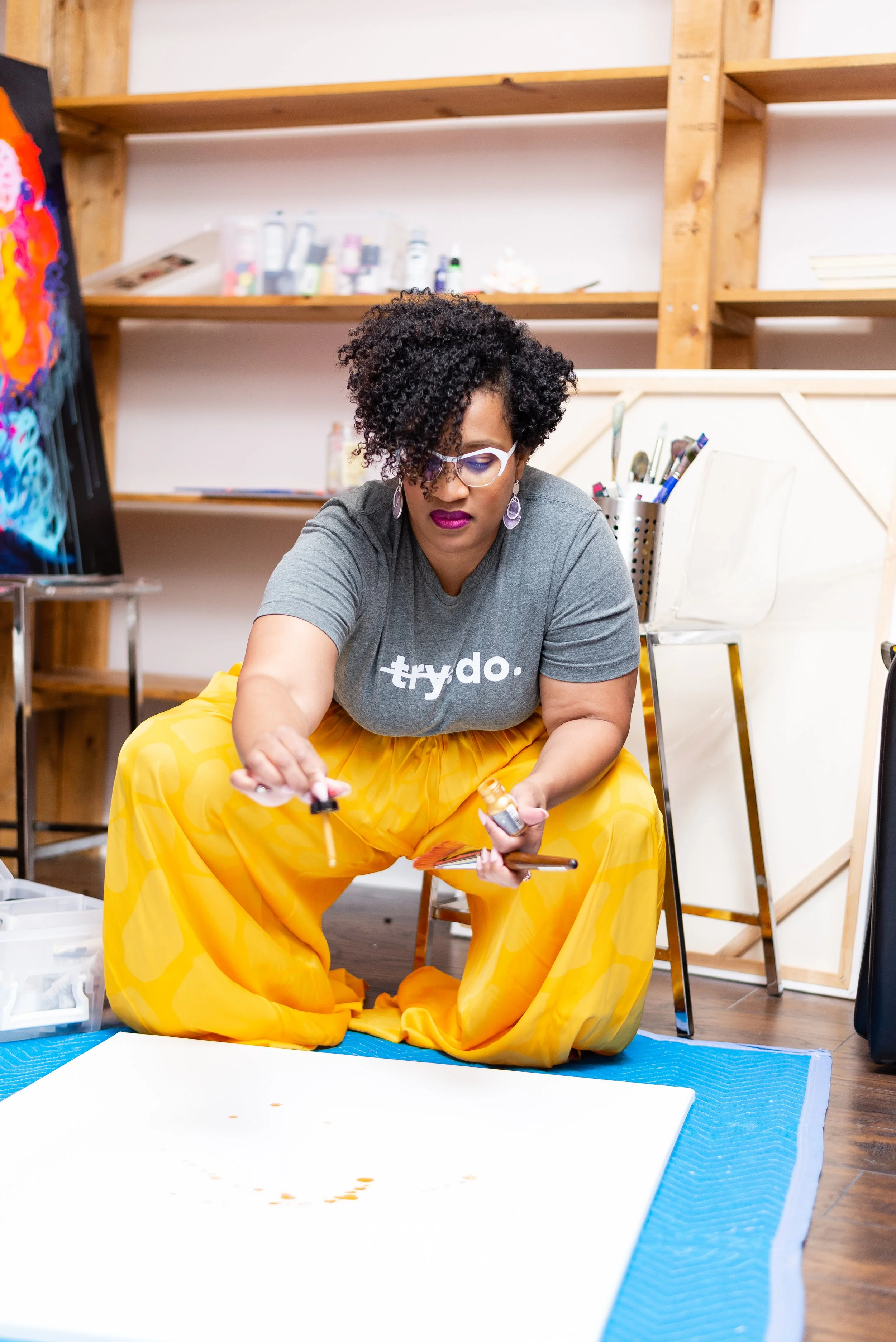 Atlanta tight artist Aeleise Jana A woman with curly hair and glasses kneeling on a blue mat, holding a small bottle and a dropper, in an art studio with shelves behind her and artwork on the wall.
