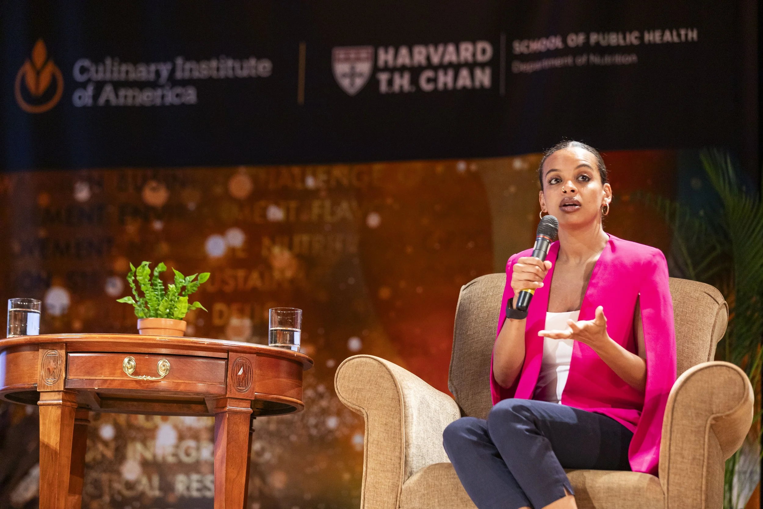 A woman in a pink blazer sitting in a beige armchair, speaking into a microphone during an event. A small wooden table with a potted plant and two glasses of water is next to her. In the background, banners with logos for the Harvard T.H. Chan School of Public Health and the Culinary Institute of America.