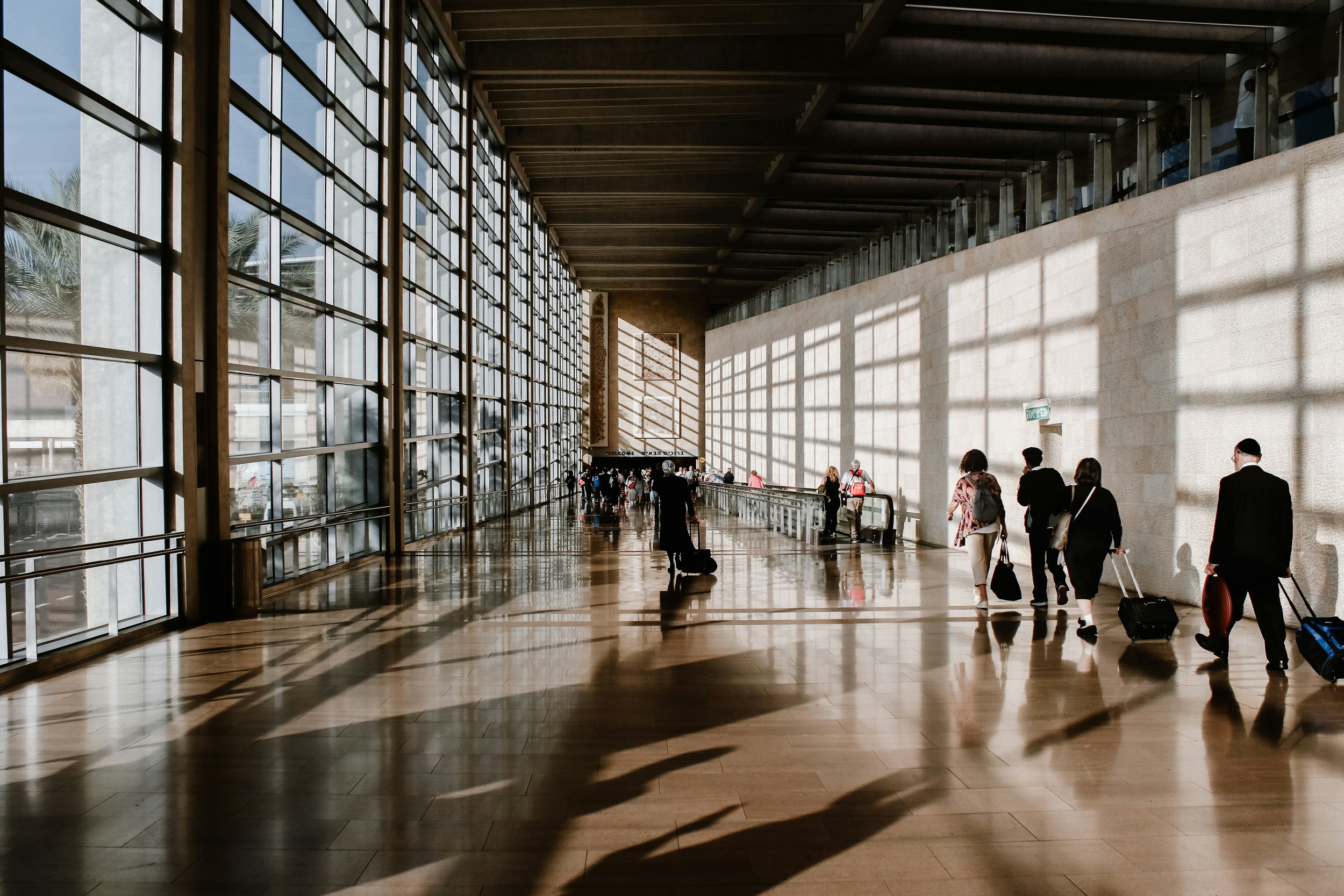 People walking through an airport terminal with large windows casting shadows on the floor.
