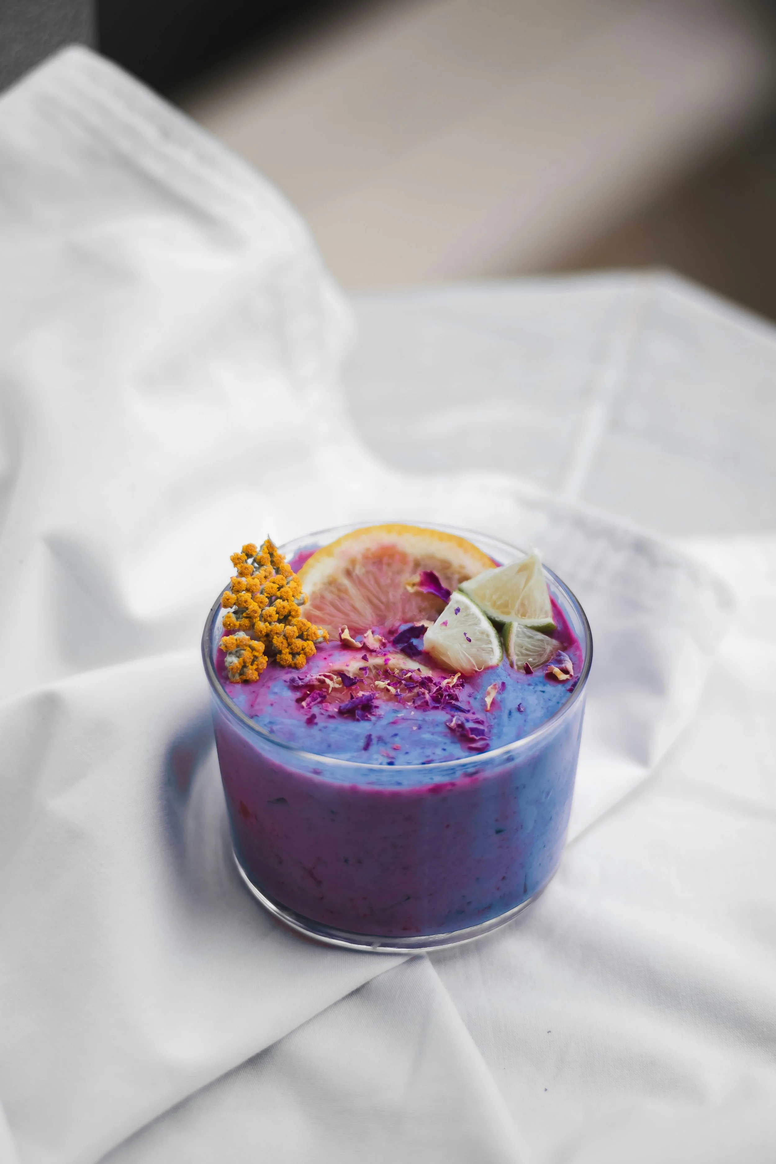 A purple smoothie in a glass jar garnished with slices of grapefruit, lime, and lemon, and sprinkled with dried flower petals and small yellow flowers, placed on a white cloth with a blurred background.