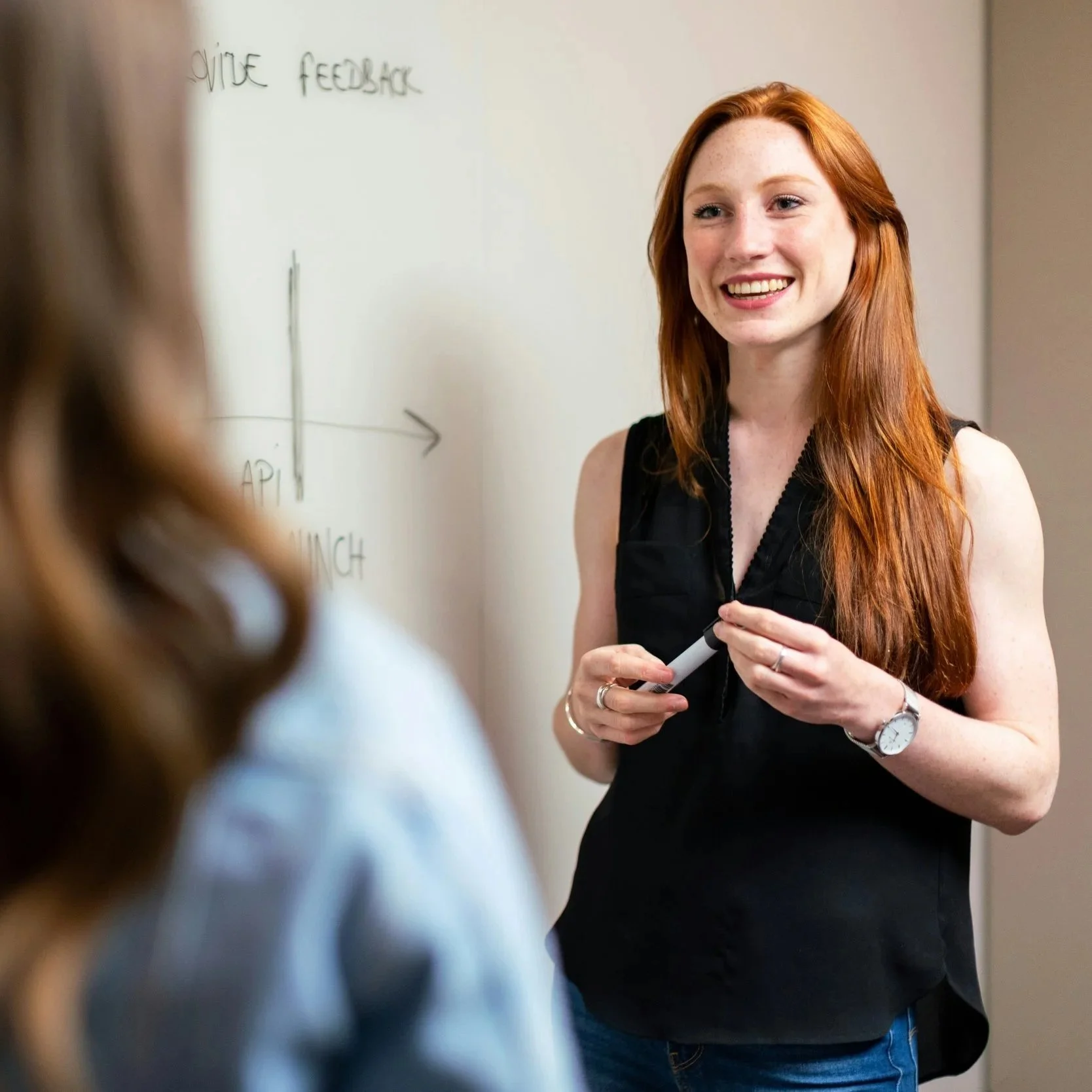 A woman with long red hair smiling and holding a marker, standing in front of a whiteboard with the words 'VOICE FEEDBACK' written at the top, engaged in a discussion with another person.