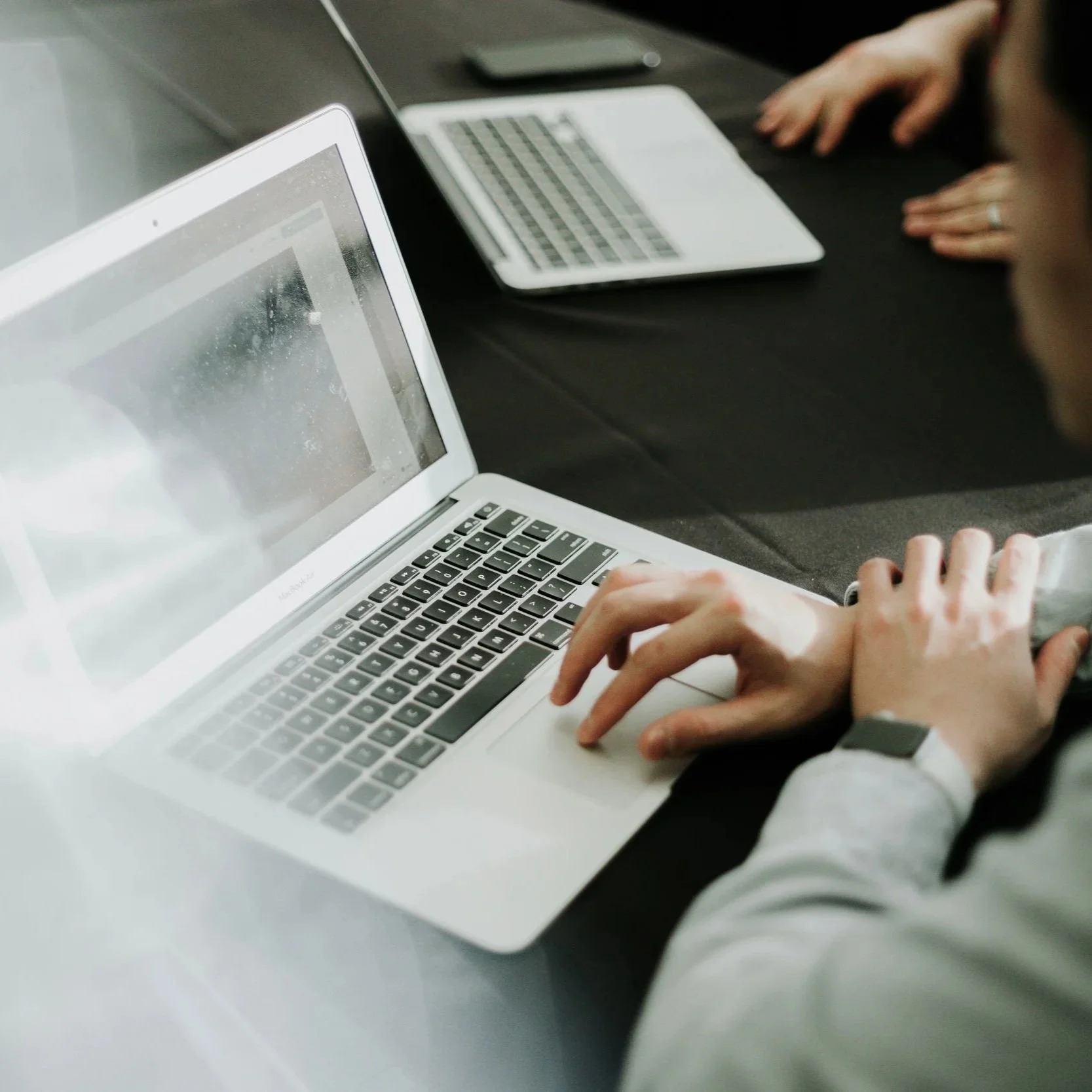 Person using a laptop at a black table, with another laptop nearby and a person's hands in the background.