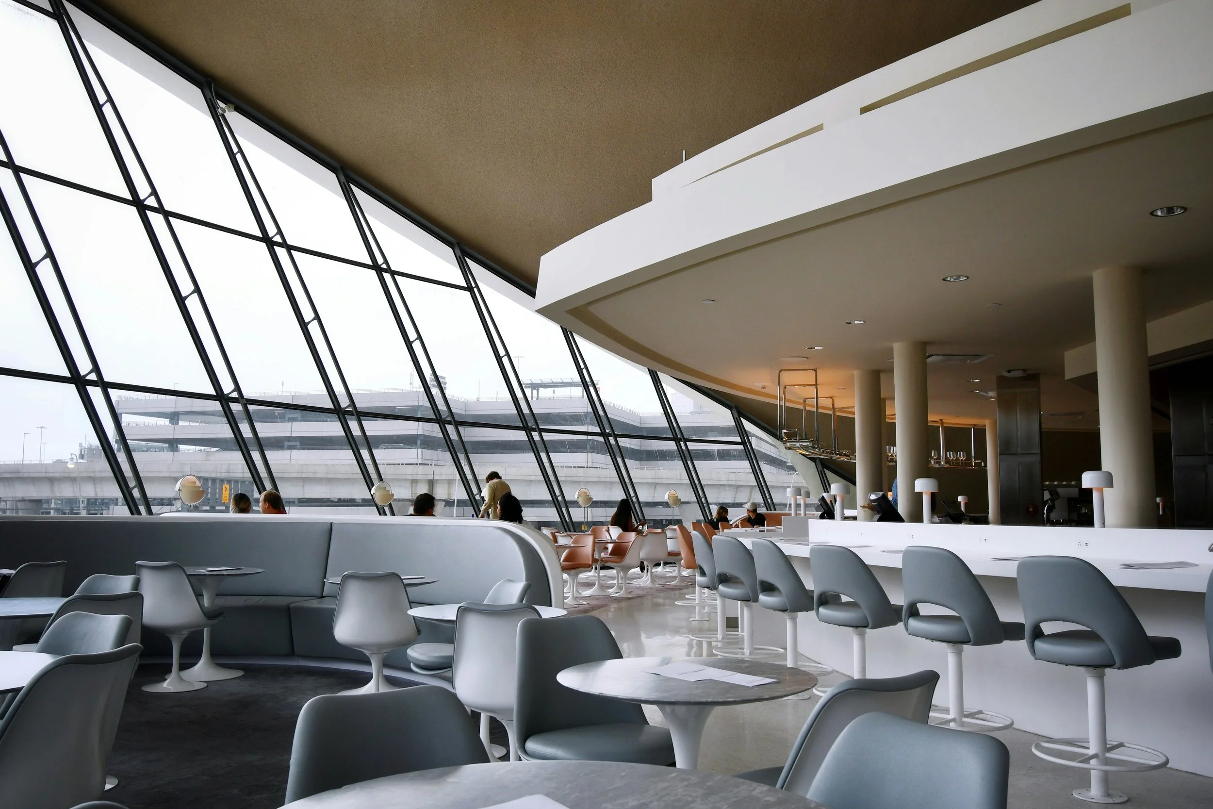 Inside an airport lounge with large glass windows, round tables, and chairs, and a bar area with barstools.