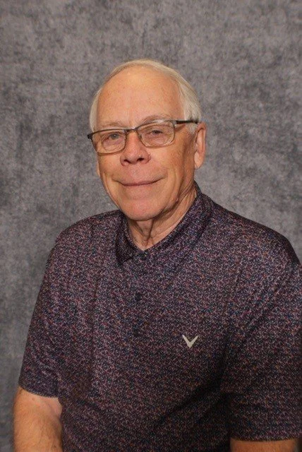 A professional headshot of an elderly man with glasses, wearing a black suit, black shirt, and a patterned silver tie, against a blue textured background.