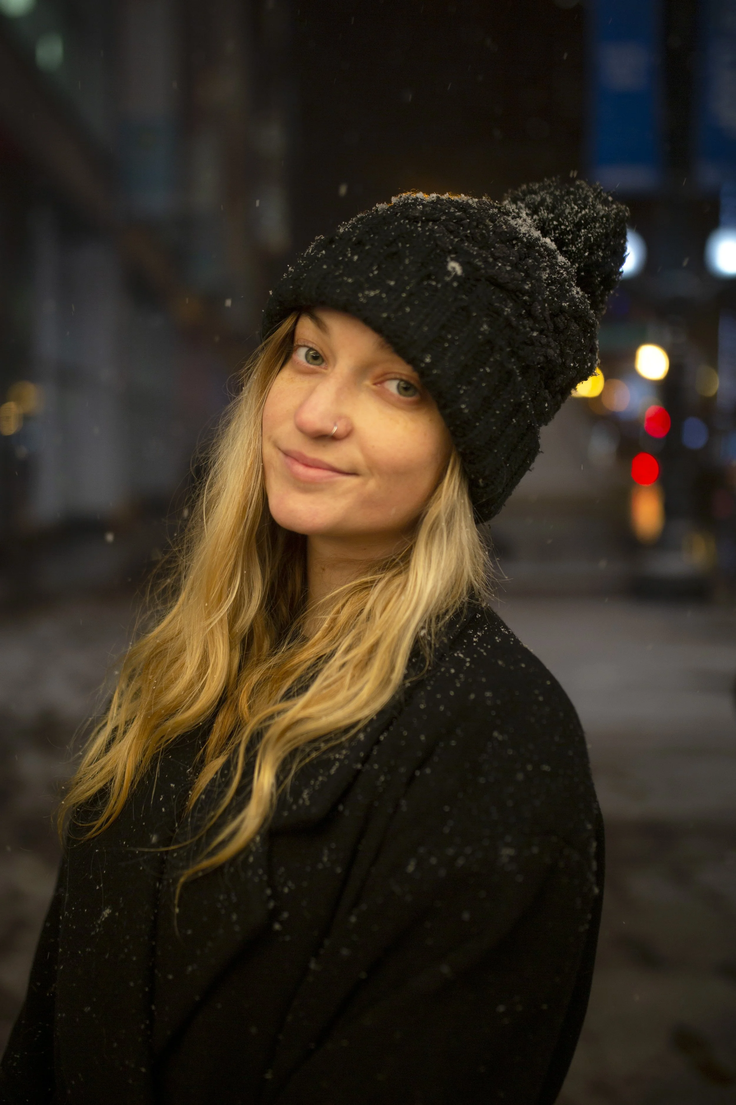 Young woman with blonde hair, wearing a beige beanie, sunglasses on her head, and a teal jacket, sits on rocks in a mountainous landscape with a forest and distant mountains in the background, sticking out her tongue playfully while holding a camera.