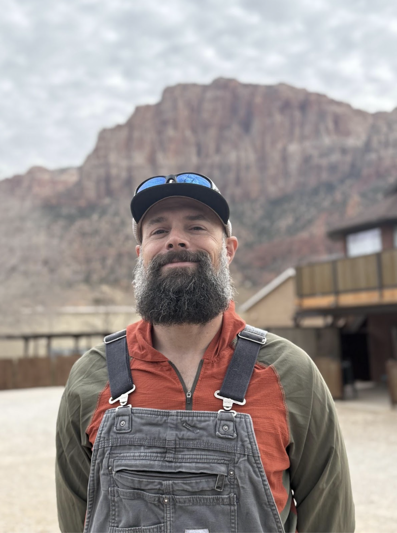A man riding a red mountain bike on a dirt trail in a wooded area, wearing a helmet, sunglasses, gloves, and casual athletic clothing.