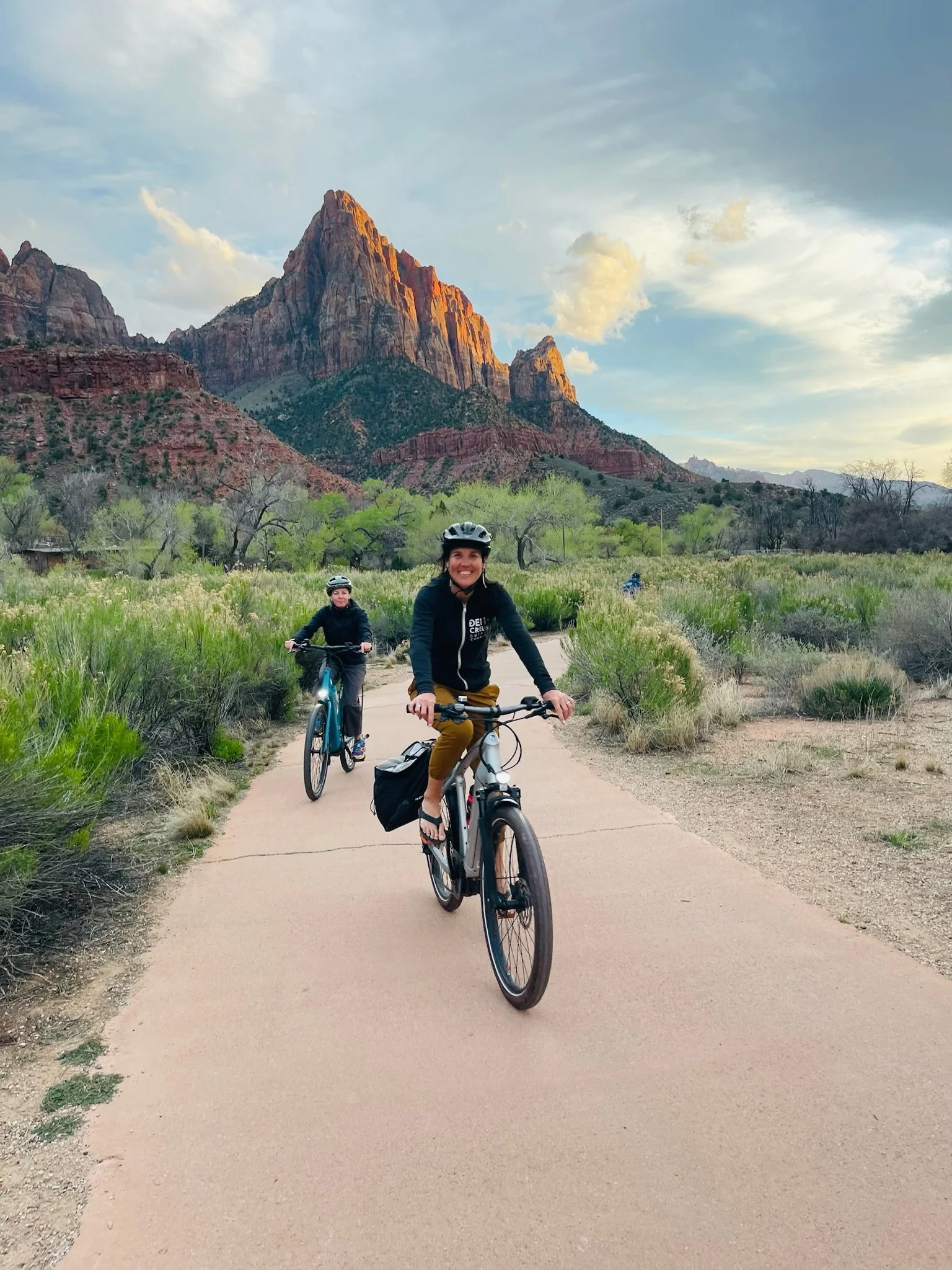 Two women riding bicycles on a trail in Zion National Park with red rock mountains in the background, green trees, and a partly cloudy sky.