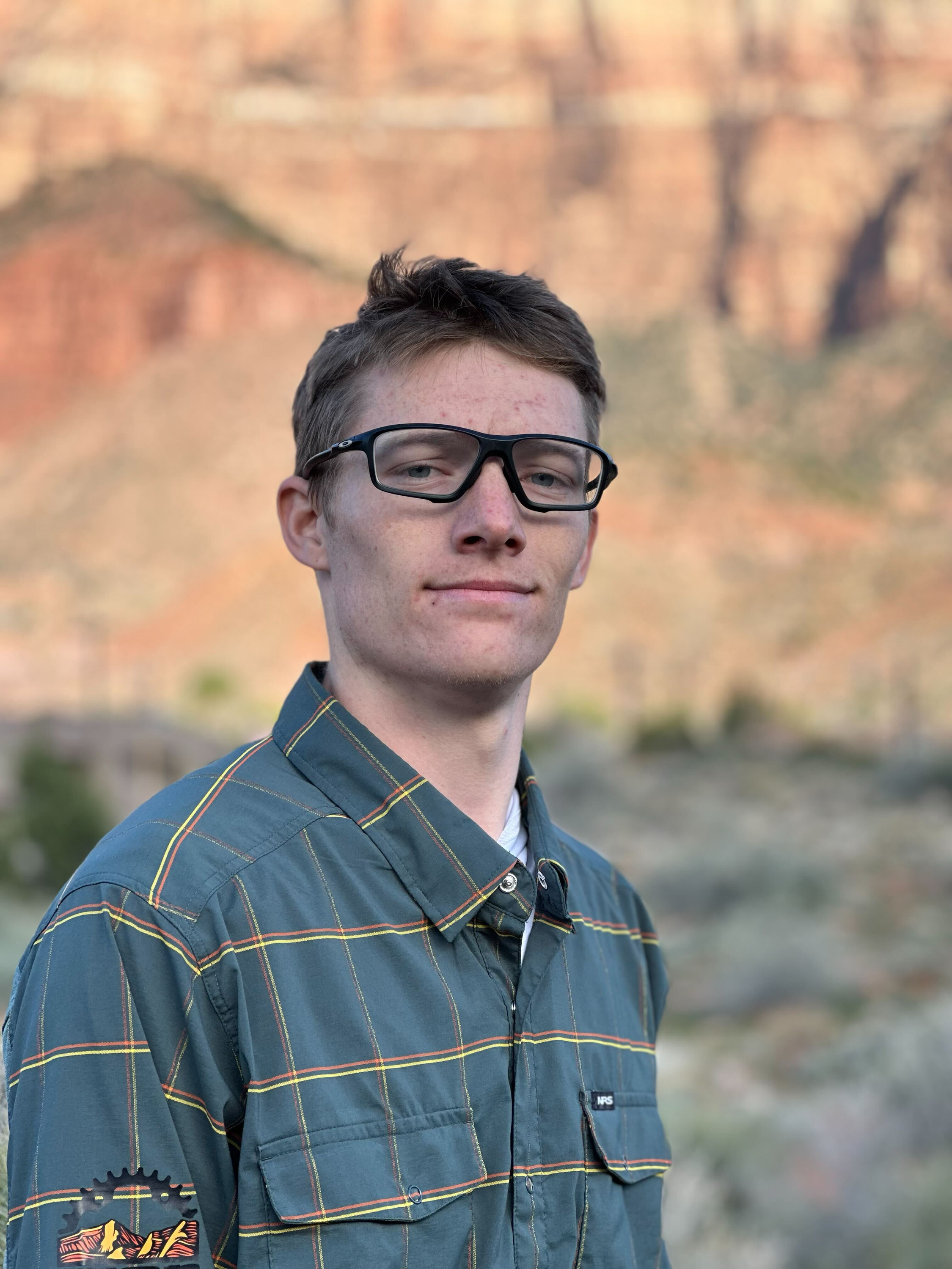 A woman wearing a bike helmet and sunglasses taking a selfie outdoors with a mountain bike and a pickup truck in the background. The terrain is desert-like with dry bushes and rocks.