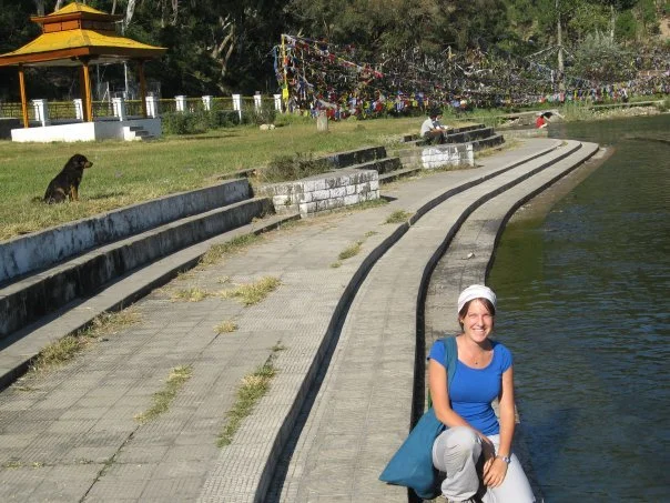 A woman sitting on a riverside walkway smiling, with a dog sitting on the grassy area behind her, and a temple with prayer flags in the background.