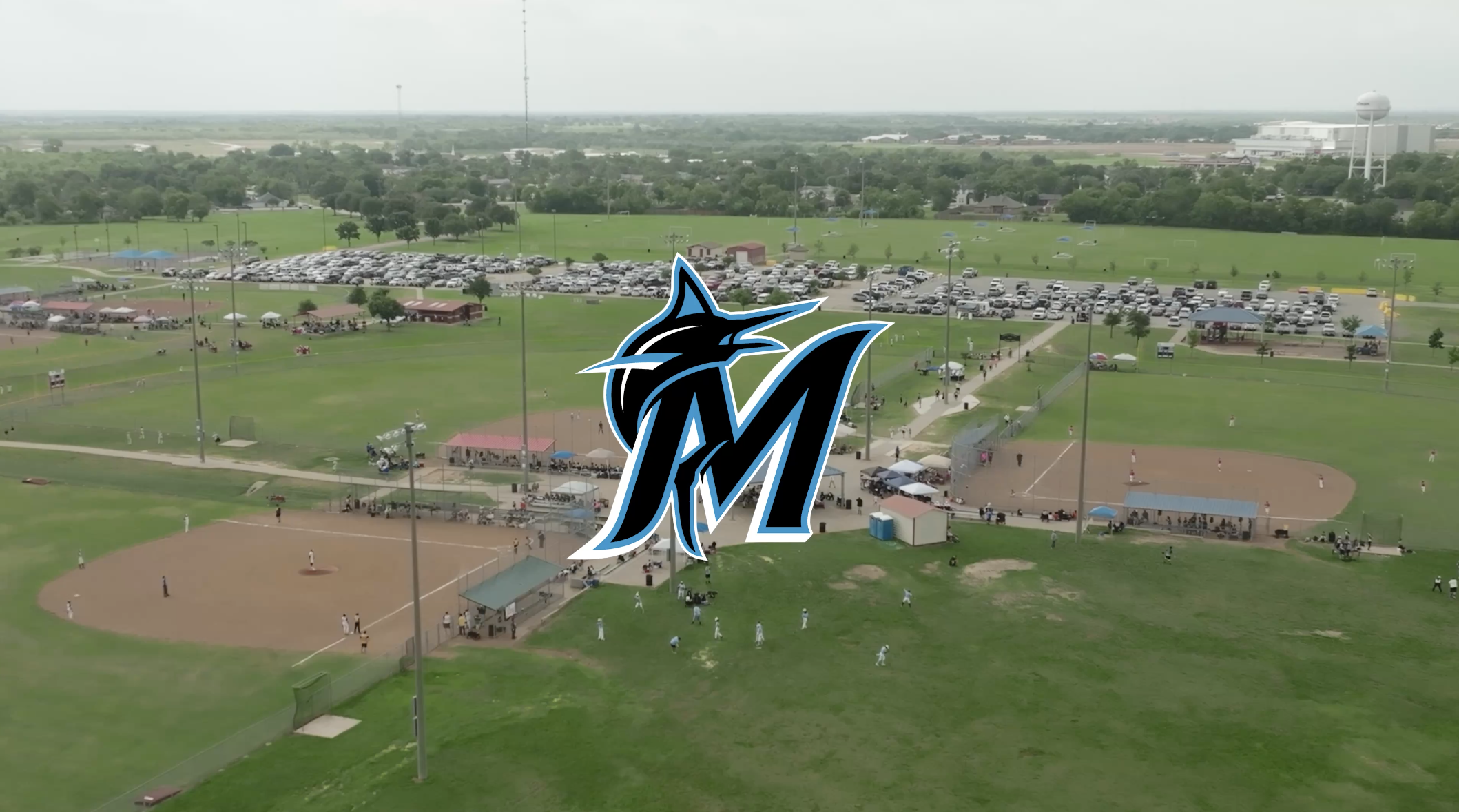 Aerial view of a baseball field with players, a parking lot, and a baseball game, with a sports team logo overlaid in the front.