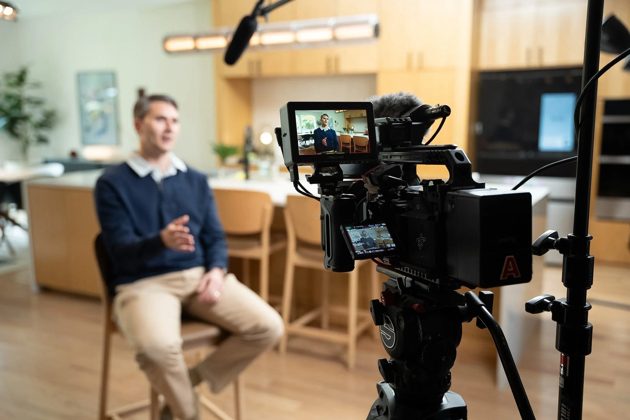 A man in a blue sweater and beige pants is sitting on a wooden chair in a room with light green walls and wooden cabinets. He is talking while being filmed by a professional camera on a tripod, with a monitor showing the shot.