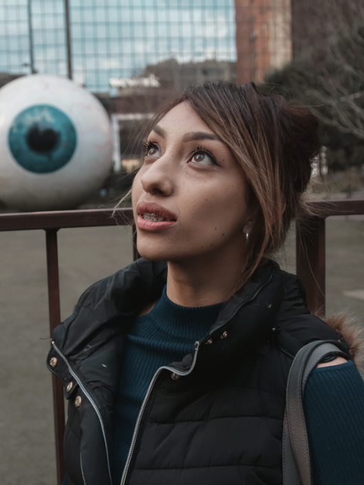 Young woman with brown hair tied in a bun, wearing a black vest and teal sweater, looking upward outdoors in front of a large photo of an eye on a building.