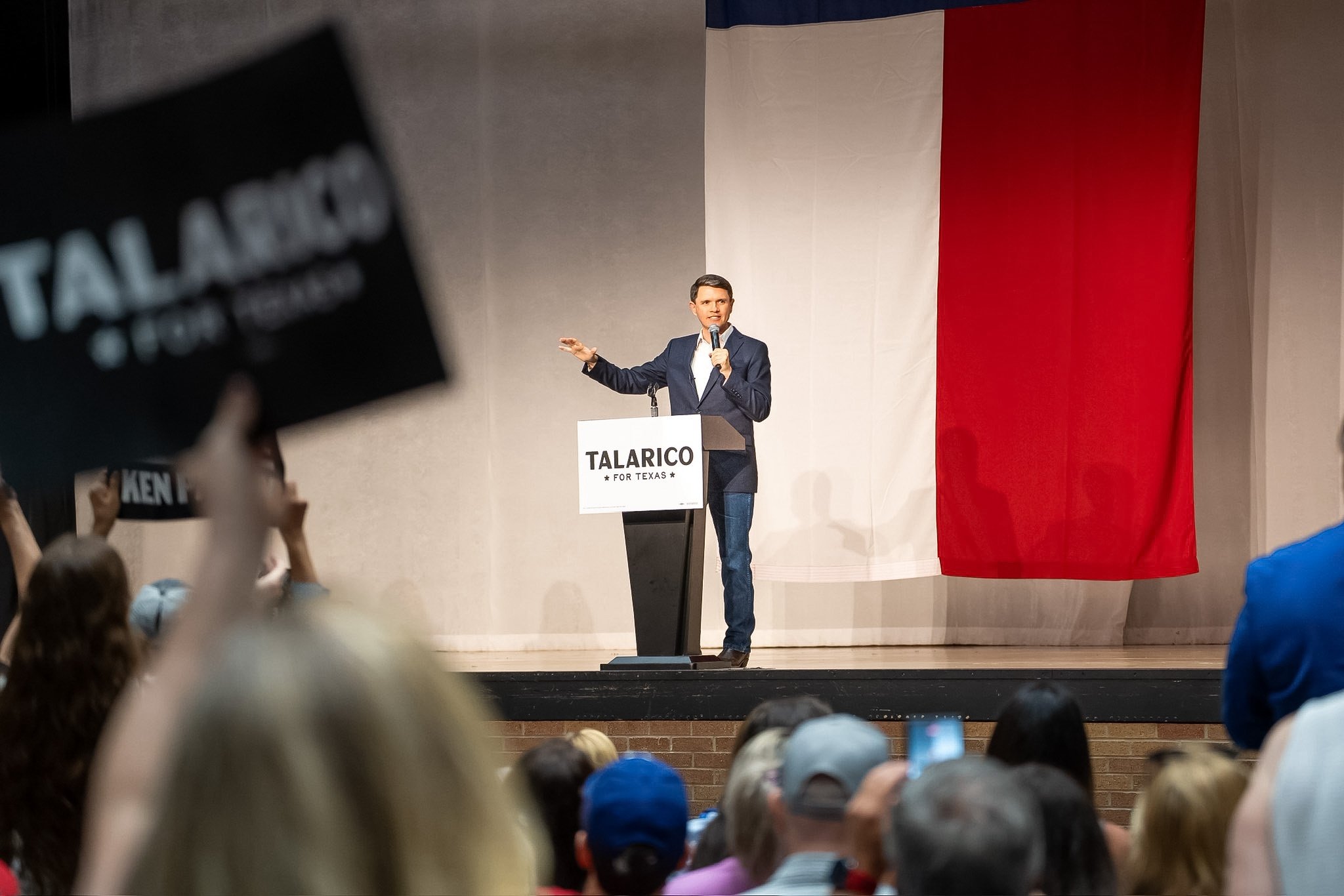 A man in a suit is speaking at a podium with a sign that reads 'TALARICO FOR TEXAS' during a political rally. The background features a large Texas flag.
