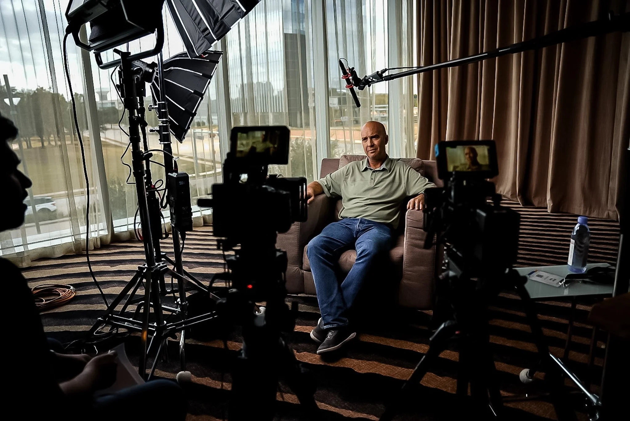 A man sitting on a brown couch in front of a large window with beige curtains, during an interview or filming. The scene is set in a room with a patterned carpet. Multiple cameras and studio lighting equipment are directed at him, capturing the interview or recording.