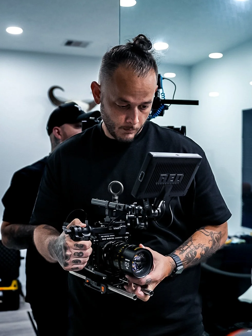 A man with tattoos on his arms holding a professional camera setup with a monitor attached, in a studio or indoor location.