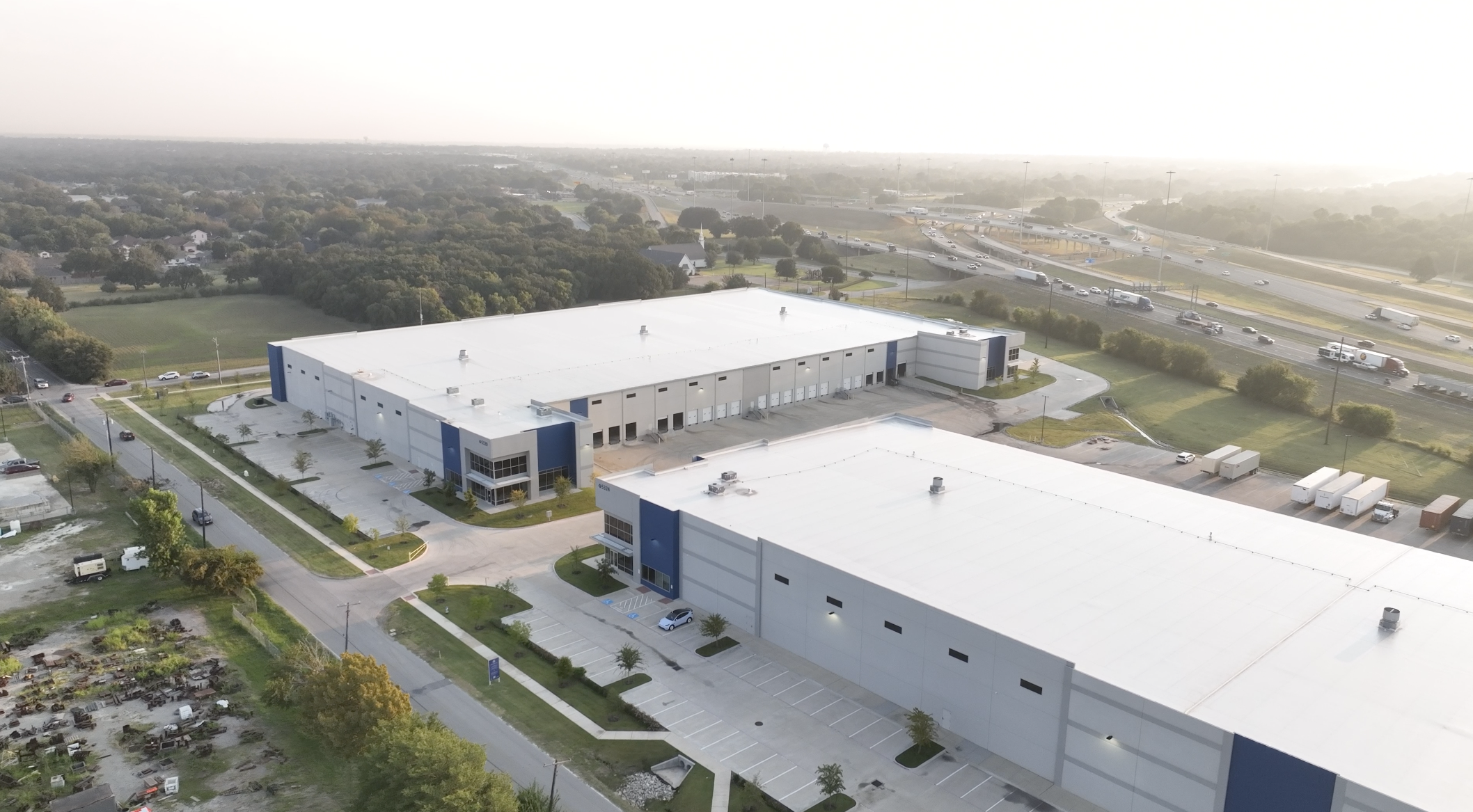 An aerial view of a large commercial warehouse with parking lots, surrounded by trees, residential houses, and a busy highway with traffic.