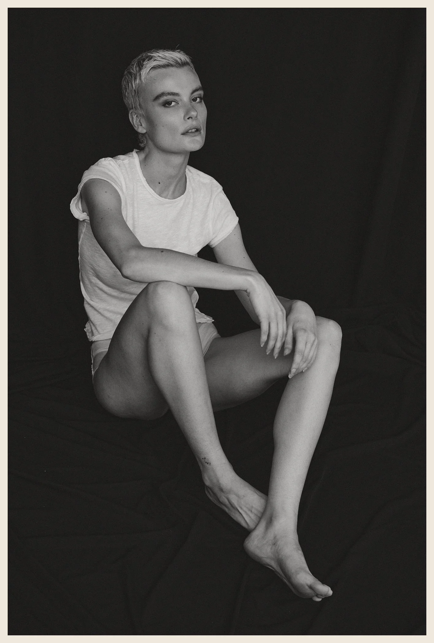 Black and white photo of a young woman with short blonde hair, wearing a white t-shirt and shorts, sitting on a dark fabric surface against a dark background, looking at the camera with a serious expression.