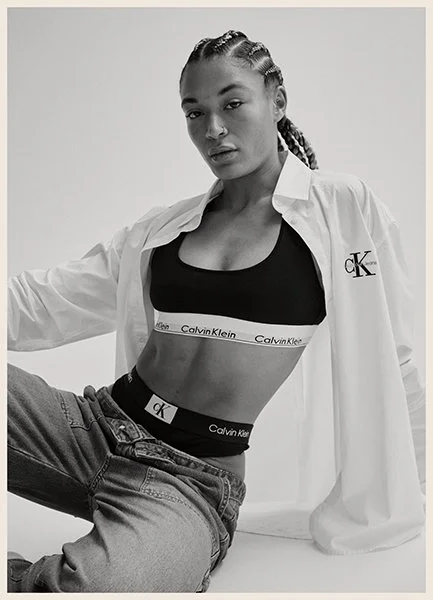 Young woman with braided hair wearing a Calvin Klein sports bra, underwear, and white shirt with CK logo, posing against a plain background.