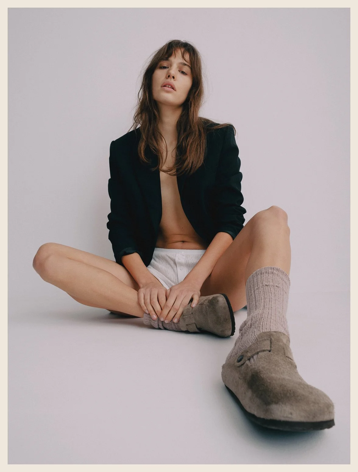 A woman sitting on the floor with her legs spread apart, wearing a black blazer, white shorts, a sock on one foot, and a beige slipper on the other, against a plain white background.