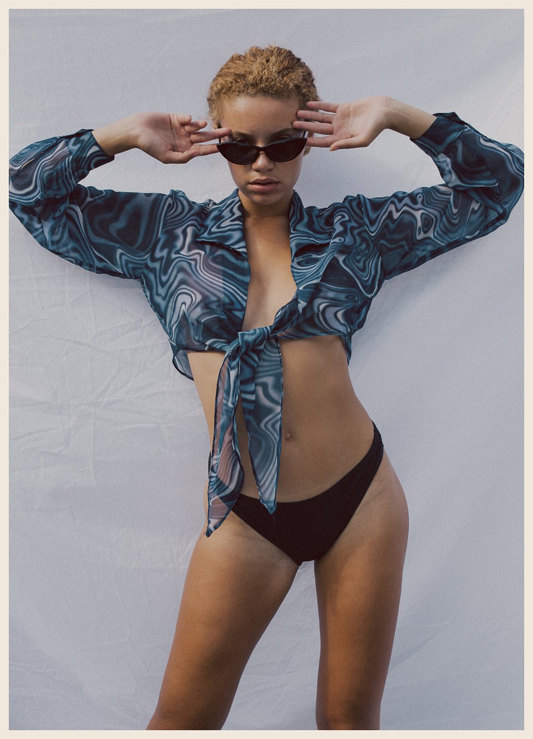 Woman wearing black bikini bottoms, a sheer blue patterned shirt tied at the front, dark sunglasses, and with short curly hair, standing against a plain background.