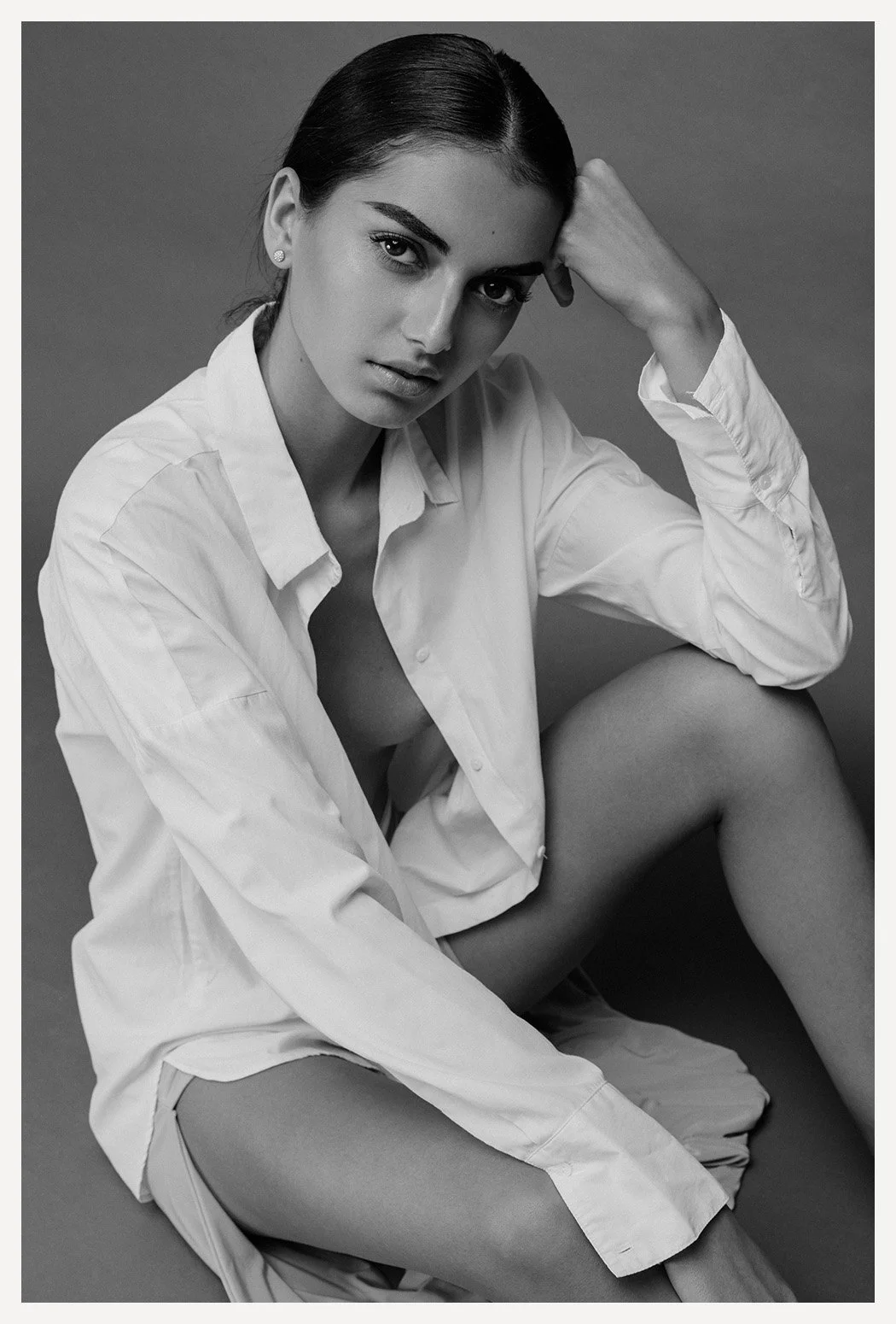 Black and white portrait of a woman wearing an unbuttoned white shirt, seated with one knee up, looking at the camera with a serious expression, against a plain gray background.