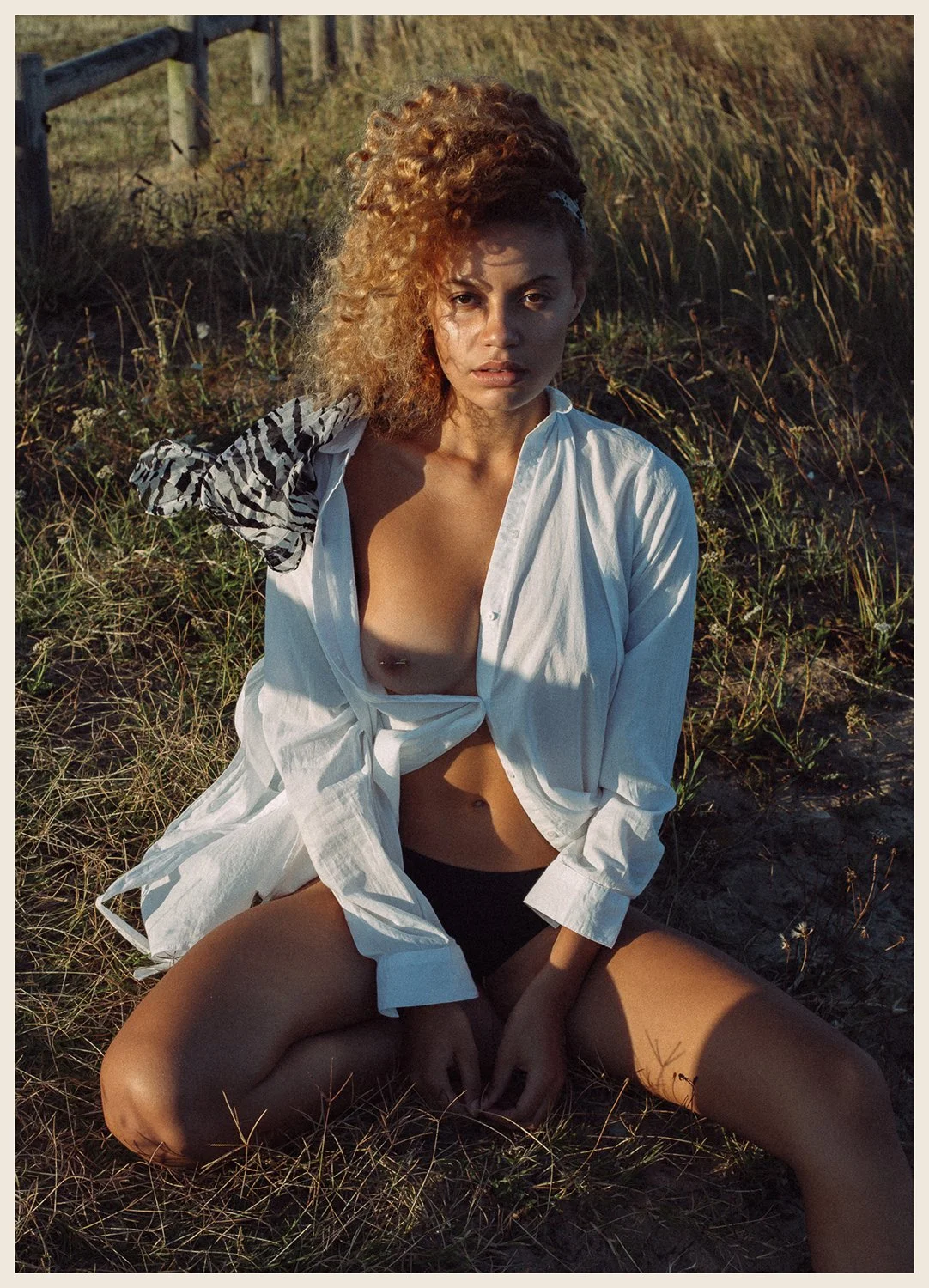 A woman with curly hair sitting on the grass in a field with tall grass and a wooden fence in the background. She is wearing a partially unbuttoned white shirt and black shorts, with a zebra-patterned scarf tied to her hair.
