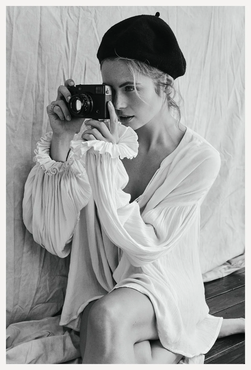 A woman sitting on the floor, wearing a black beret, taking a black and white photograph with a vintage camera. She has curly hair, a white dress with pleated, voluminous sleeves, and is partially in shadow, with a crumpled backdrop behind her.