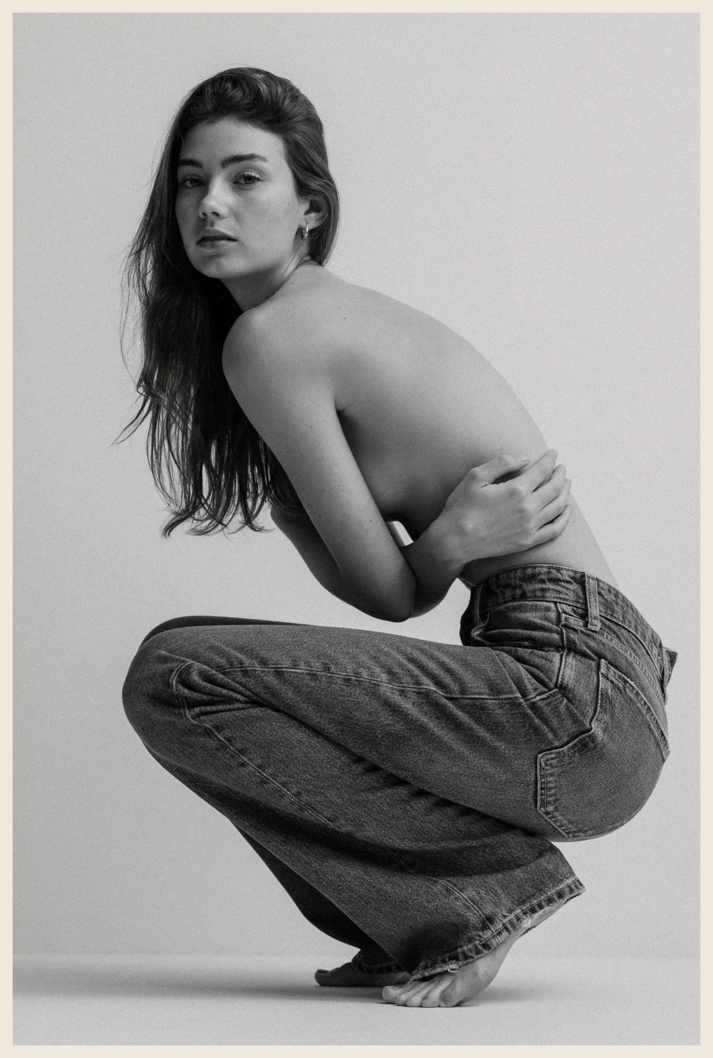 Black and white photo of a topless woman with long hair, wearing jeans, crouching with arms crossed over her chest, facing slightly to the left, against a plain light background.