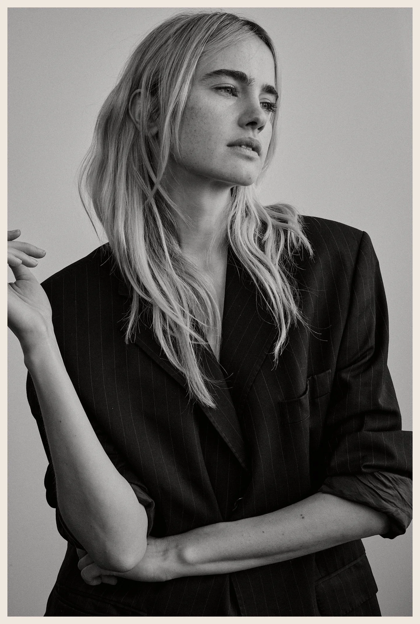 Black and white portrait of a woman with long blonde hair wearing a pinstripe blazer, gazing to the side with a contemplative expression, her arms crossed.