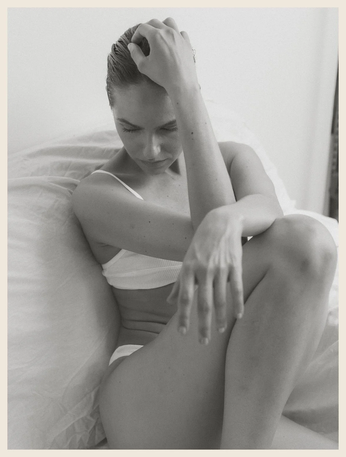 A black and white photo of a woman sitting on a bed, wearing a tank top and underwear, with her eyes closed, holding her head with one hand and extending one arm and leg, in a contemplative pose.
