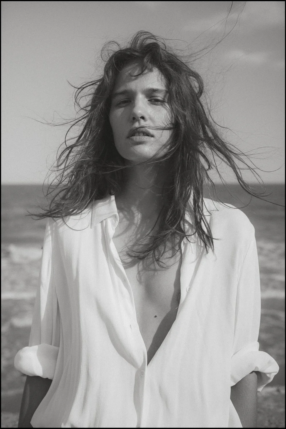 A black and white photograph of a woman with messy hair standing on a beach, wearing an open white shirt.