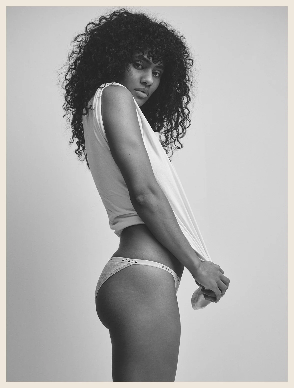 Black and white photo of a woman with curly hair, wearing a sleeveless top and underwear, standing against a plain background.