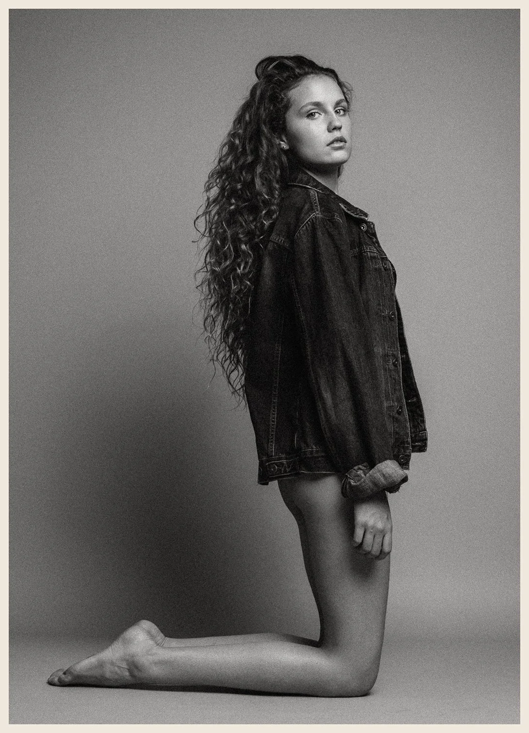 A black and white photo of a woman with long curly hair, kneeling on the floor in a studio. She is wearing a denim jacket and is topless underneath, with one hand resting by her side and the other hand clenched into a fist. Her gaze is directed towar