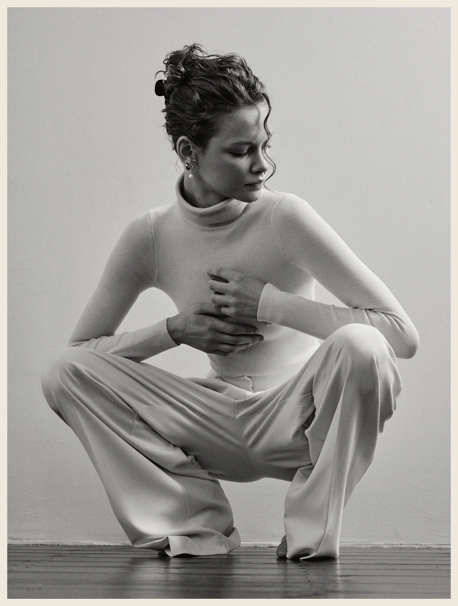 A black-and-white photo of a woman in a turtleneck and wide pants, squatting on the floor with one hand on her chest and looking to her left.