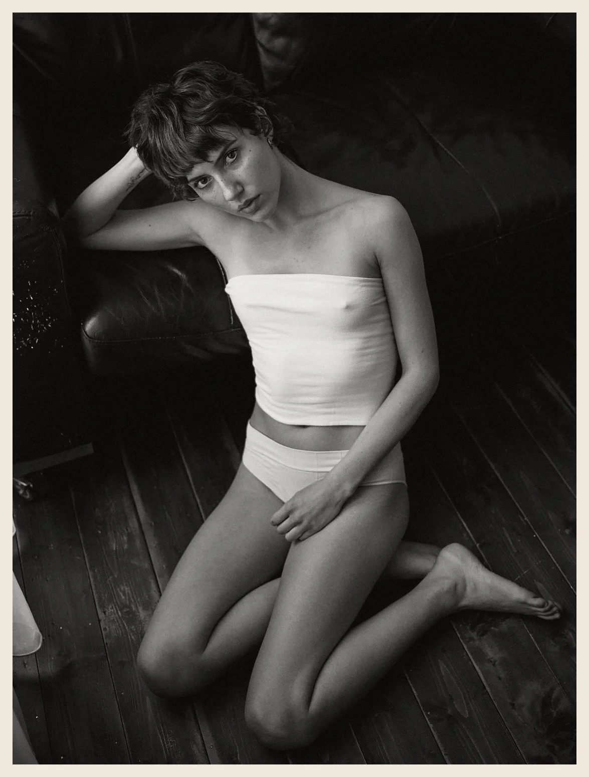 A young woman with short dark hair, sitting on her knees on a wooden floor, leaning against a dark leather sofa, wearing a strapless white top and matching underwear, looking into the camera.