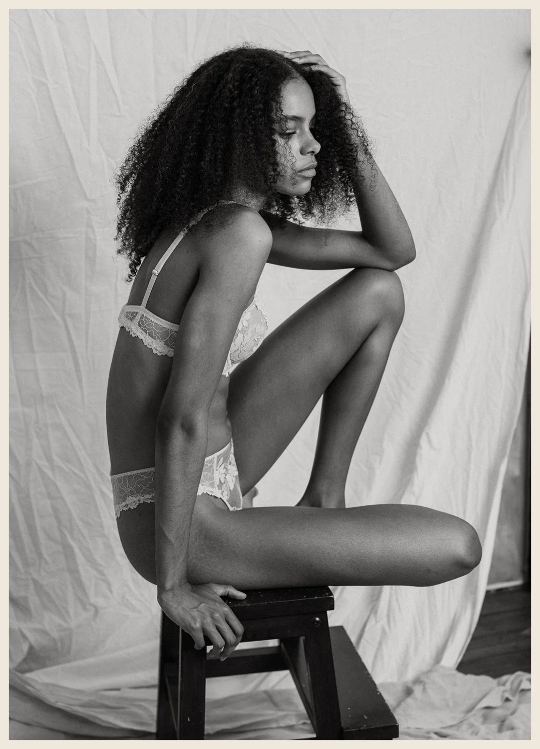 A black and white photo of a woman with curly hair sitting on a stool, wearing lace underwear, with one leg bent and resting on the stool, and her hand on her head, against a plain backdrop.