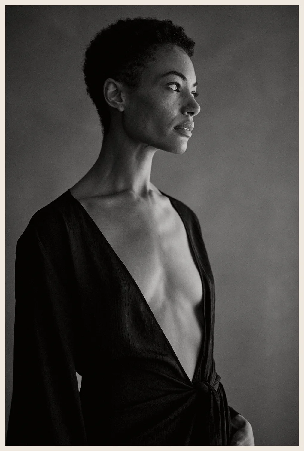 Black and white portrait of a woman with short, curly hair, looking to her right with a calm expression, wearing a deep V-neck top.
