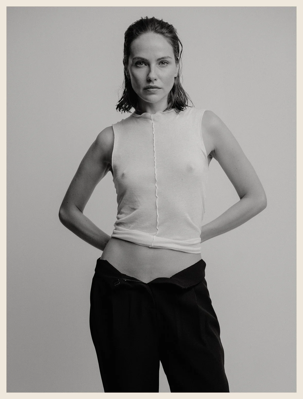 A black-and-white photo of a woman with shoulder-length hair, wearing a sleeveless top and pants, posing with her hands behind her back.