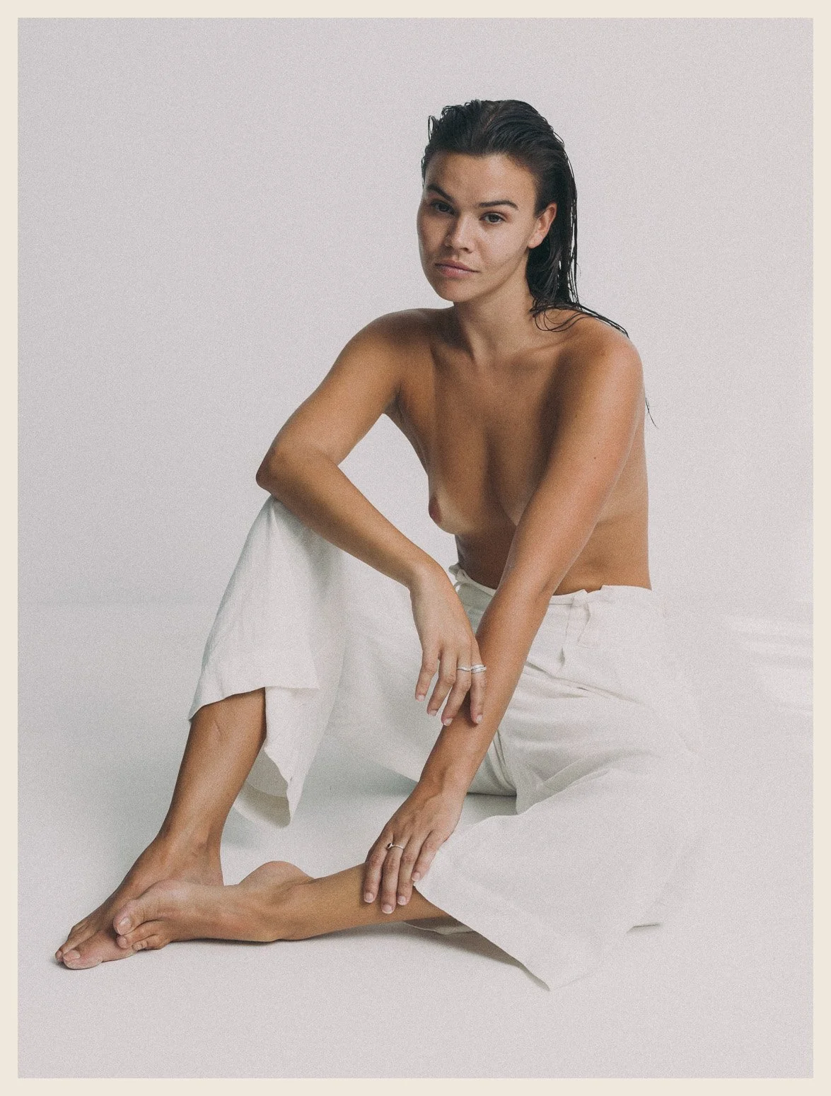 A topless woman with long dark hair seated on the floor against a plain white background, wearing white pants.
