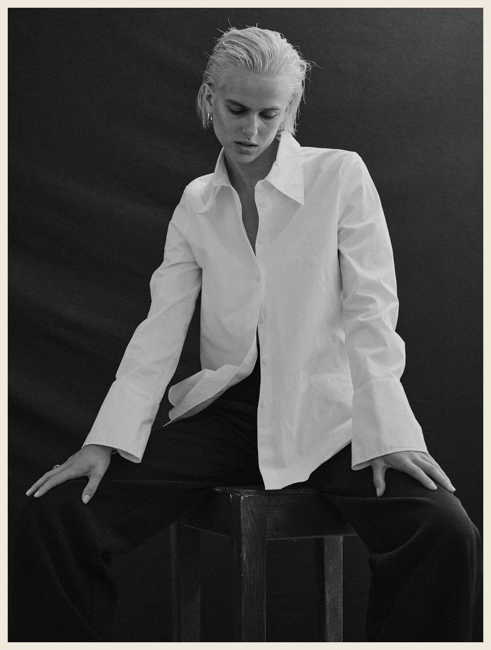 A woman with short, platinum blonde hair sitting on a wooden stool against a dark background, wearing a white button-up shirt and black pants.