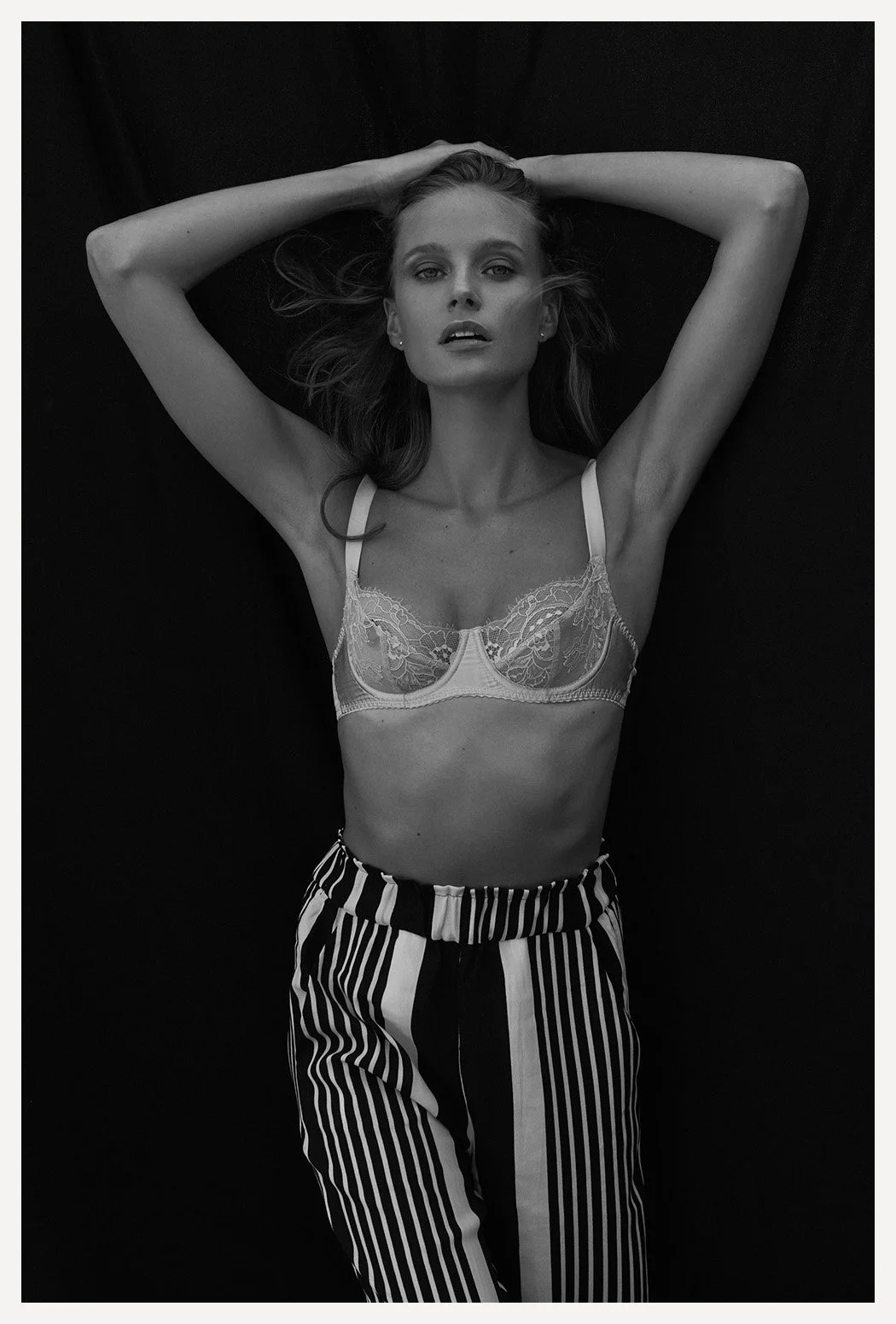 A woman with long hair lying on her back on a dark surface, wearing a lace bra and striped pants, looking at the camera.