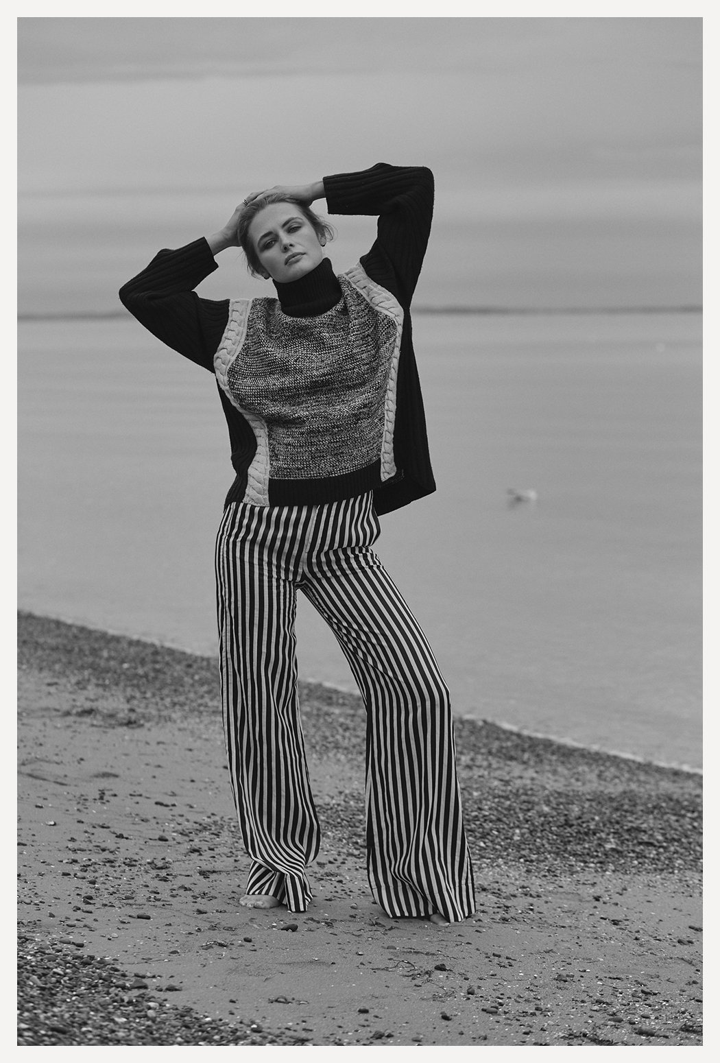 Woman standing on a beach with water in the background, wearing a patterned sweater, striped wide-leg pants, and a jacket.
