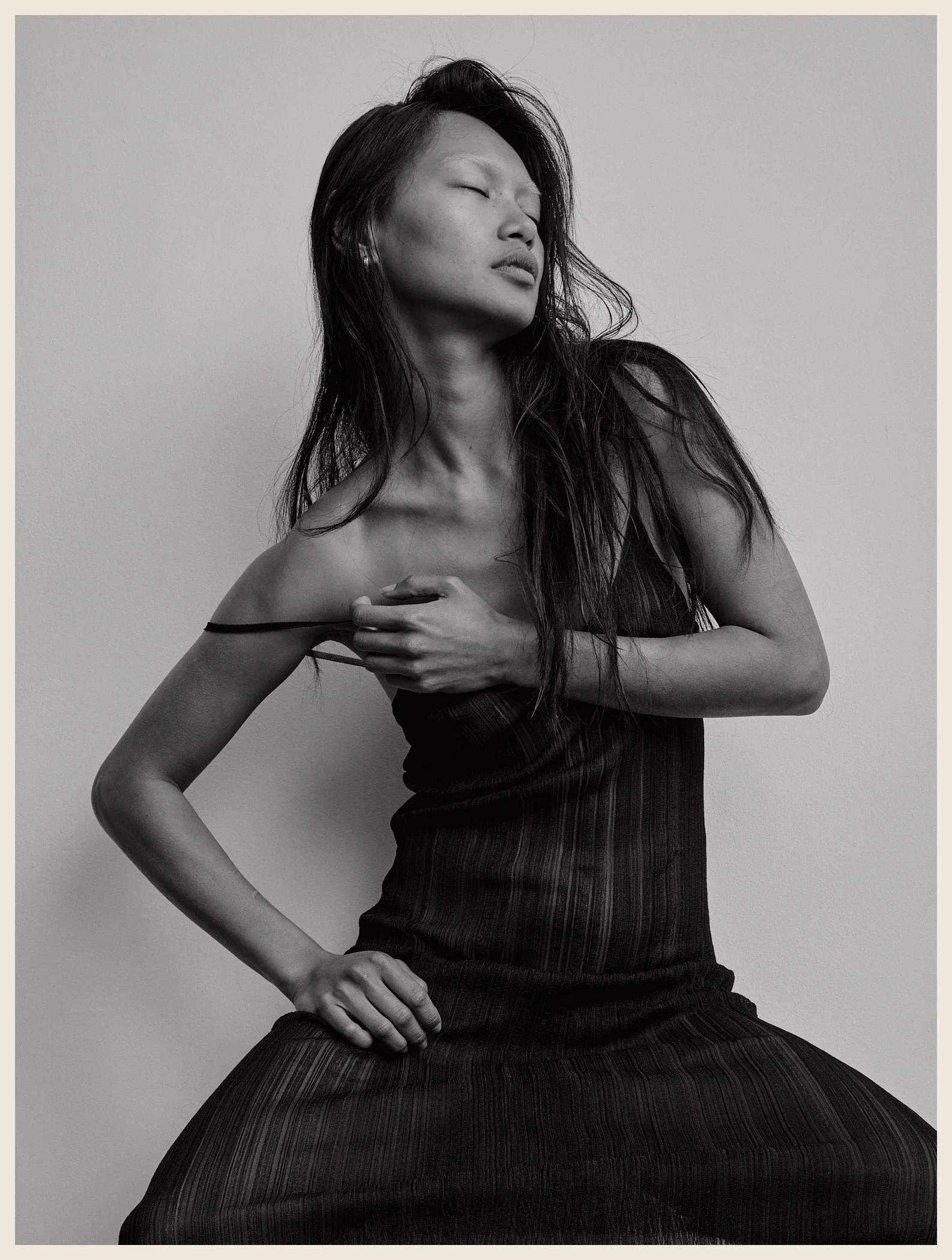 Black and white photo of a woman with long hair, wearing a dress, with eyes closed, one hand on her chest, and the other on her waist.
