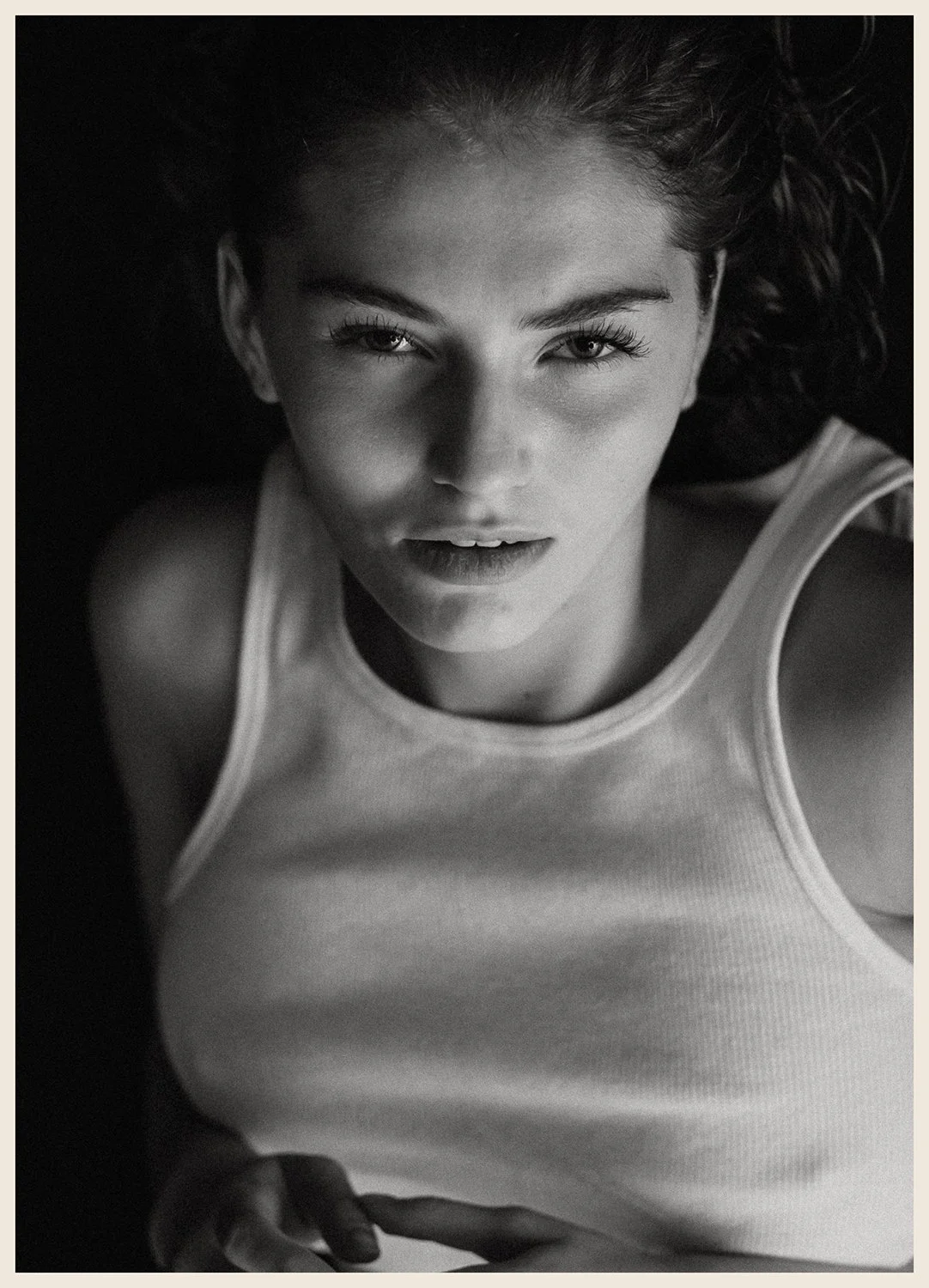 Close-up black and white portrait of a woman with curly hair, wearing a sleeveless top, looking directly at the camera.