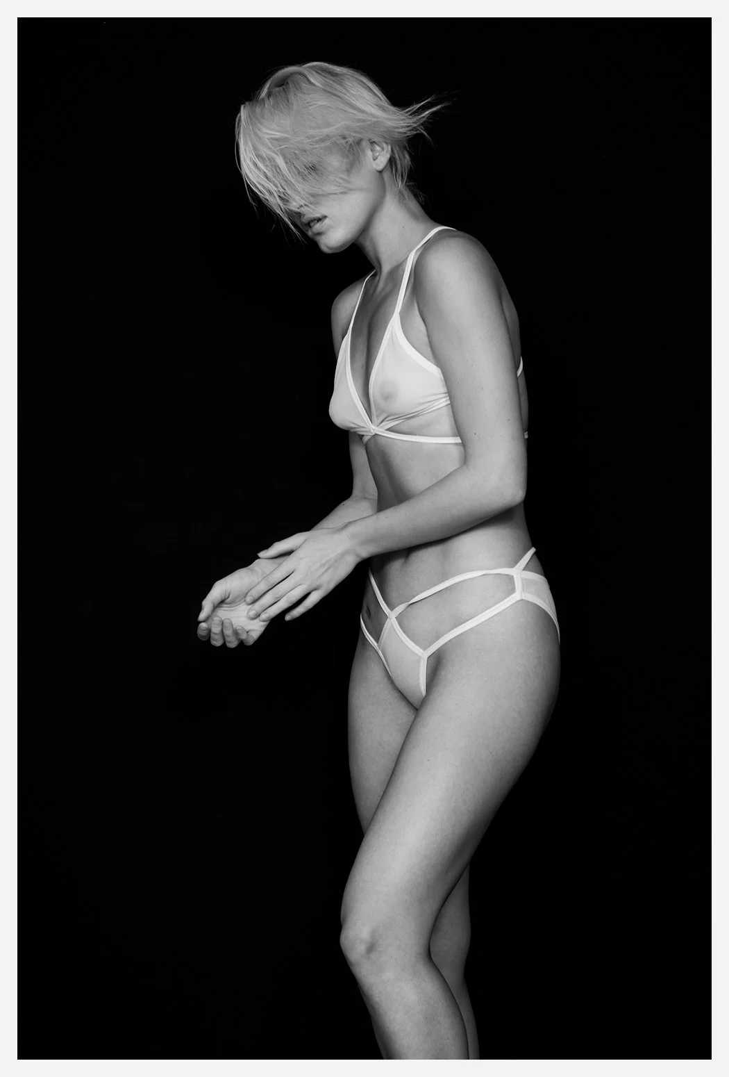 A black and white photo of a woman in lingerie with her hair covering her face, against a black background.