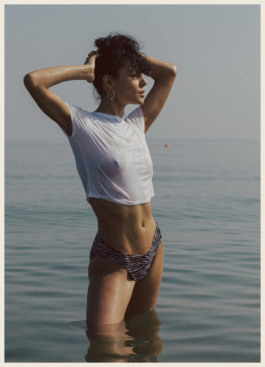 A woman standing in shallow water at the beach, wearing a wet white t-shirt and zebra-print bikini bottoms, with her hands in her hair, looking towards the horizon.