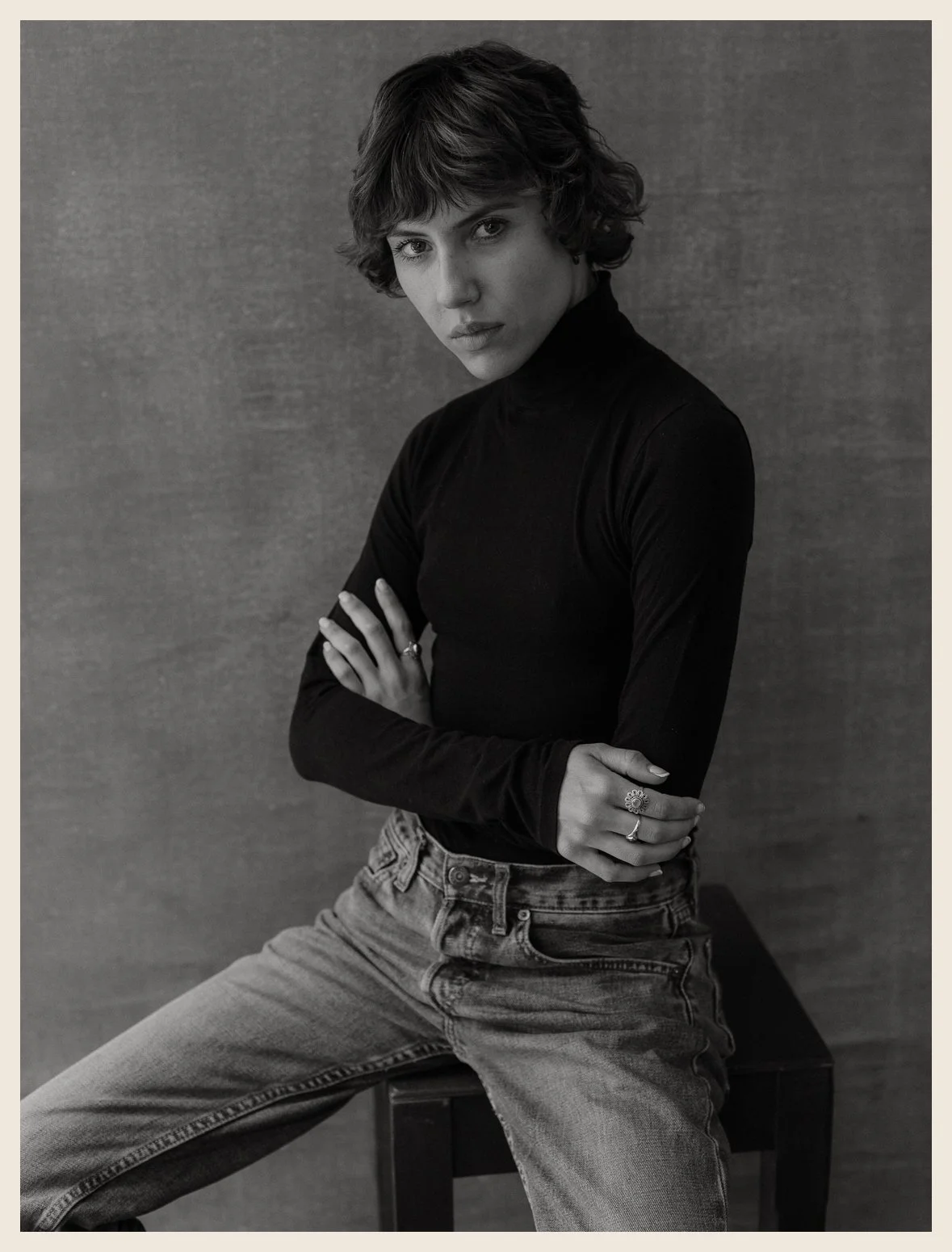 Black and white portrait of a young woman with short, wavy hair, wearing a black turtleneck and high-waisted jeans, seated on a stool against a plain backdrop, looking directly at the camera with arms crossed.
