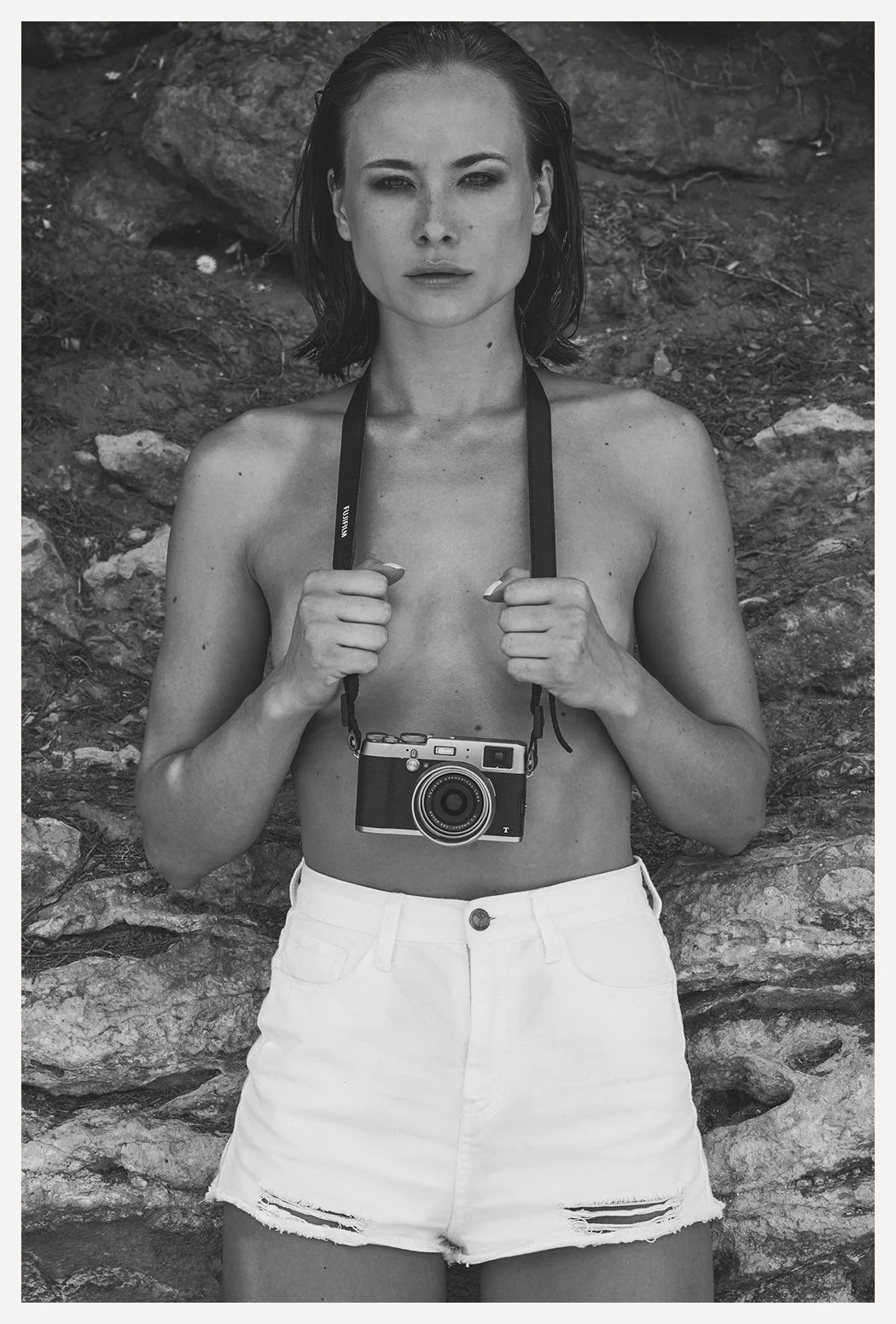 Black and white photo of a topless woman holding a camera around her neck, standing in front of a rocky background.