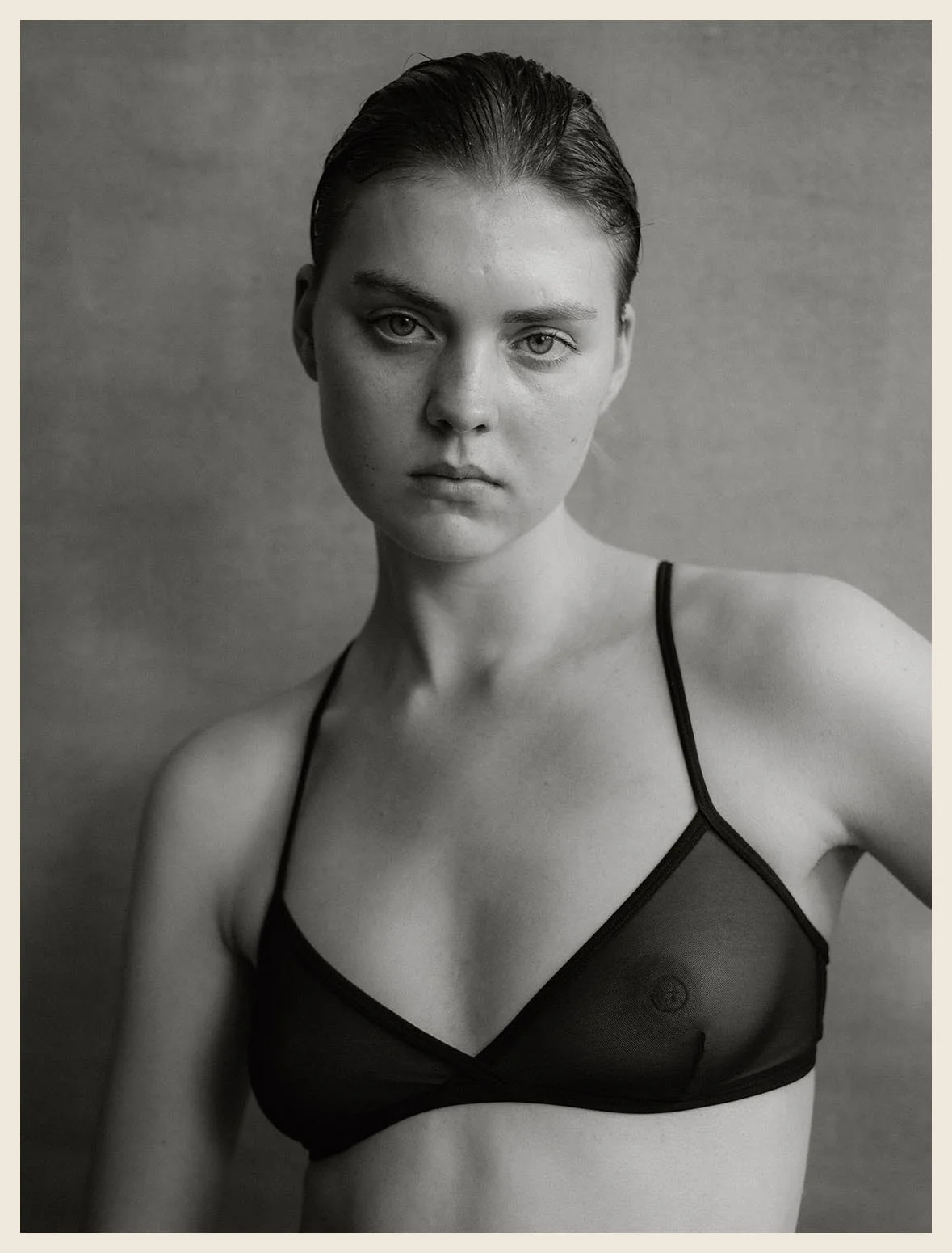 Black and white portrait of a woman with wet hair, wearing a sheer black bra, looking directly at the camera with a serious expression.